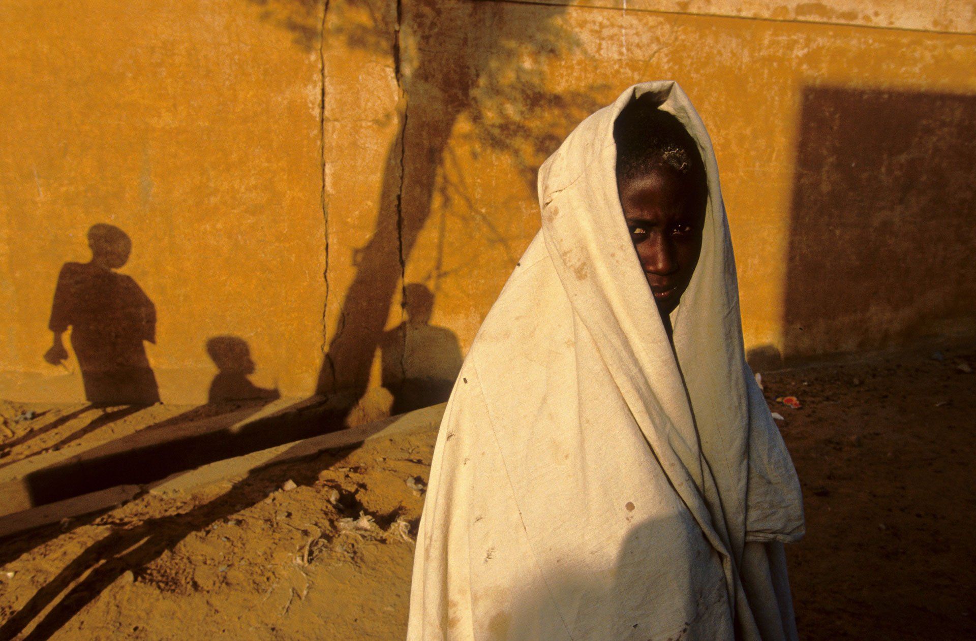 A young girl wrapped in a white blanket is standing in front of a yellow wall.