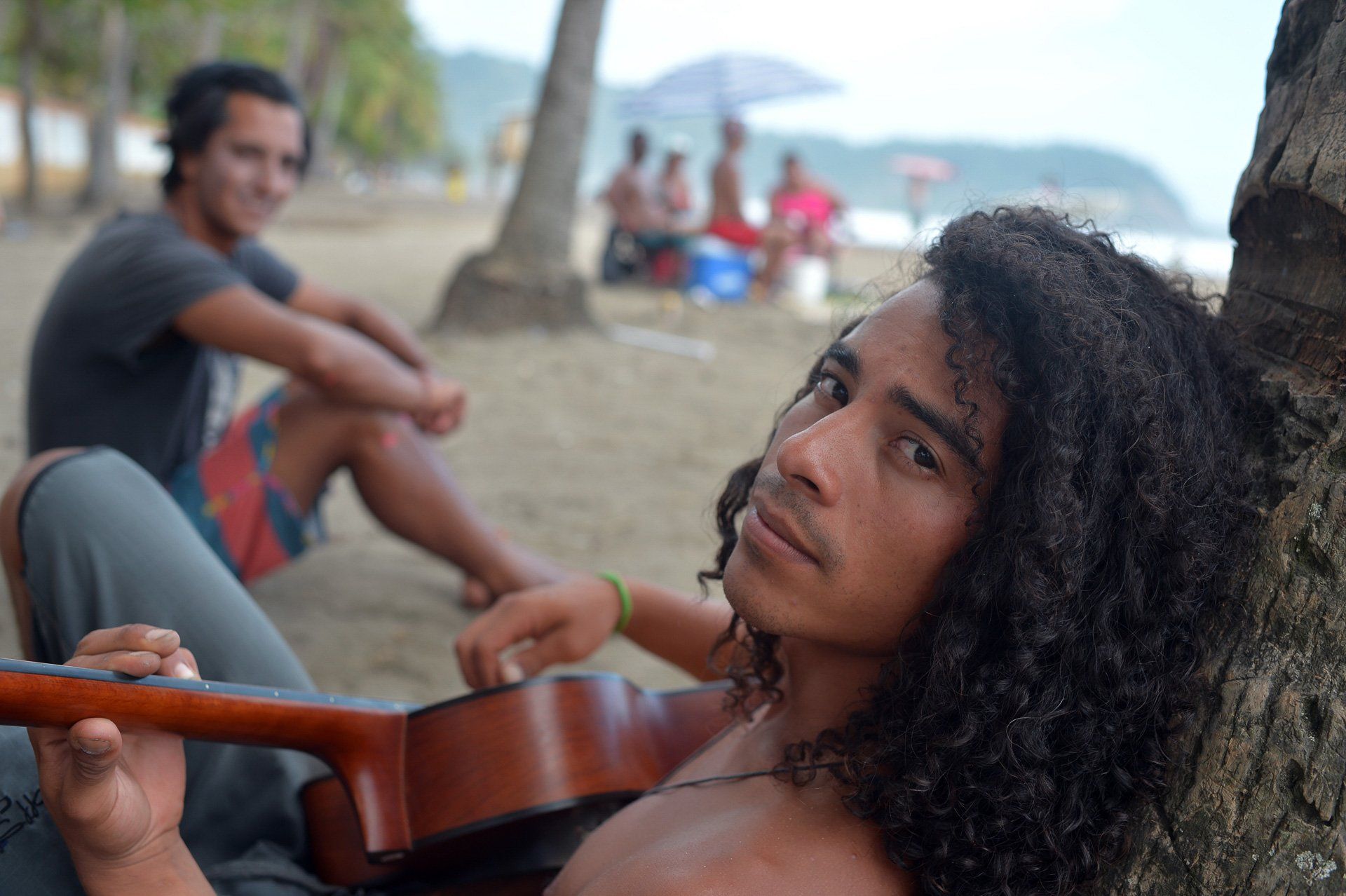 A man with long curly hair is leaning against a tree holding a guitar.