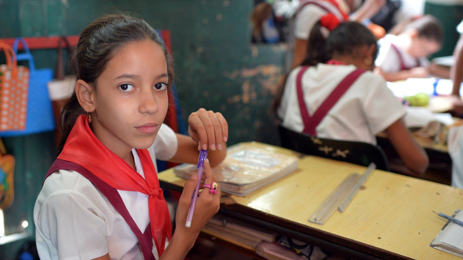 A young girl is sitting at a desk in a classroom holding a pen.