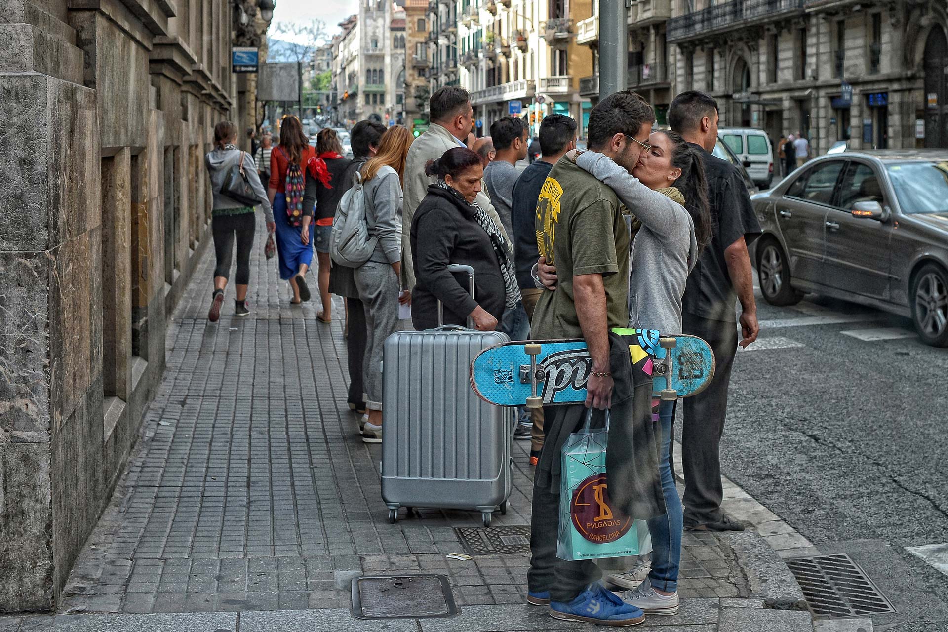 A group of people are standing on a sidewalk holding suitcases and skateboards.