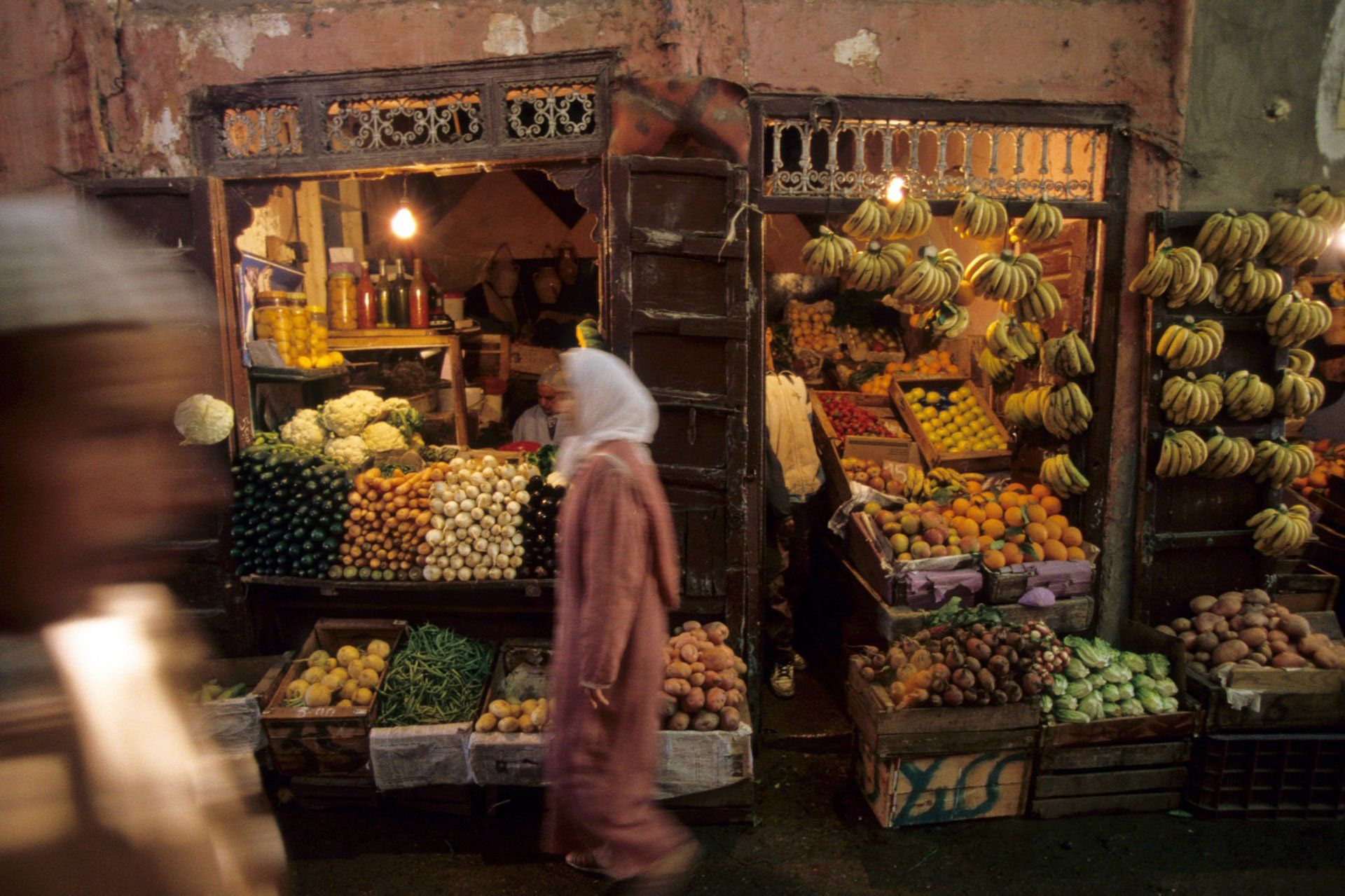 A woman is walking past a fruit and vegetable stand