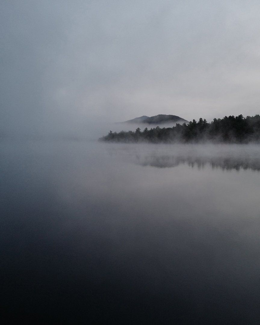 A foggy lake with mountains in the background and trees in the foreground.
