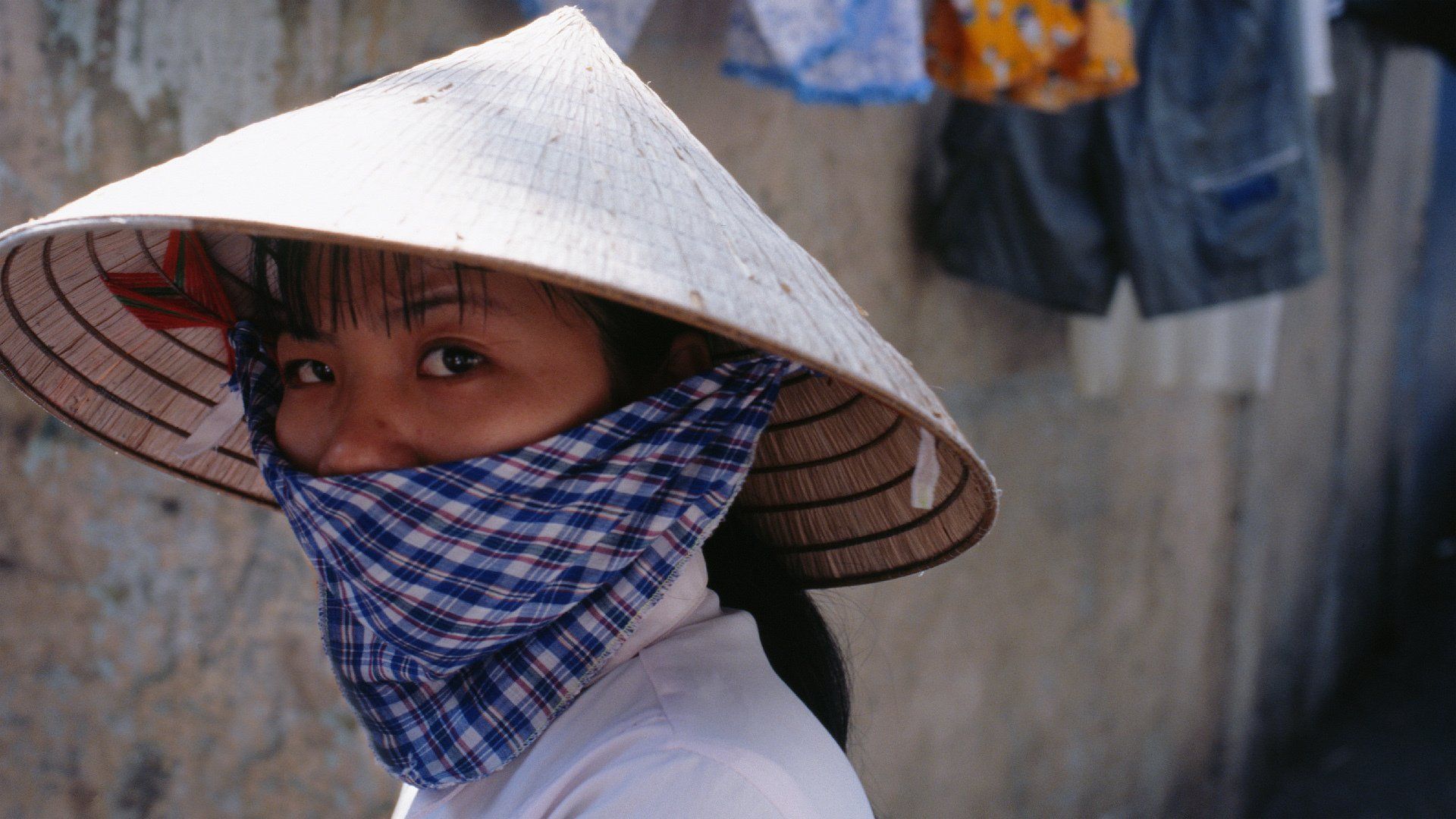 A woman wearing a conical hat and covering her face with a plaid scarf.