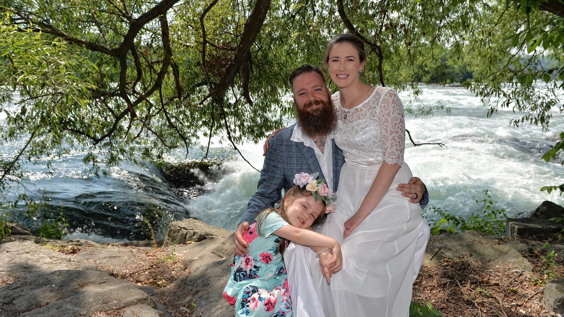 A bride and groom are posing for a picture with their daughter in front of a waterfall.