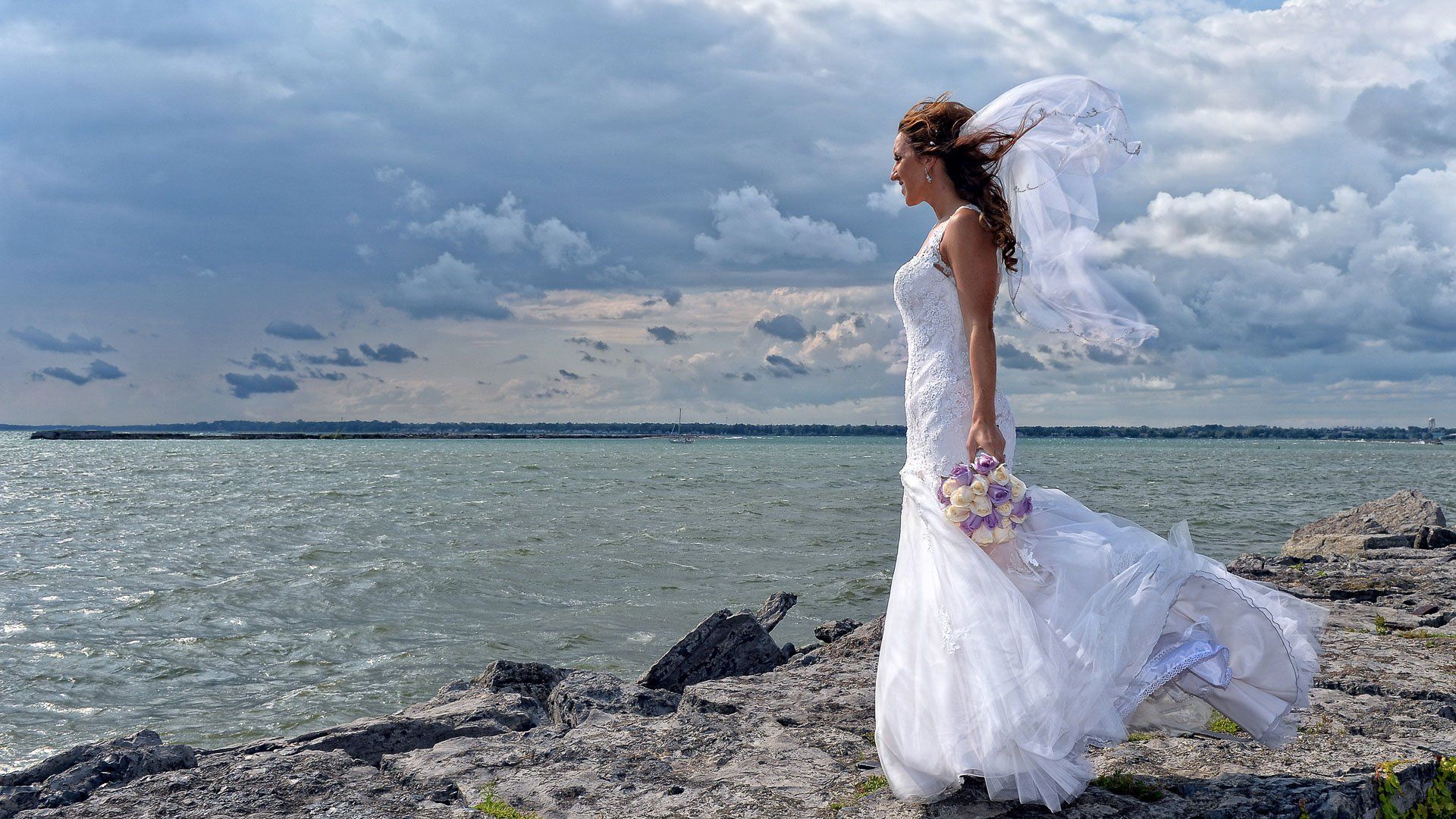 A bride in a wedding dress and veil is standing on a rocky beach near the ocean.