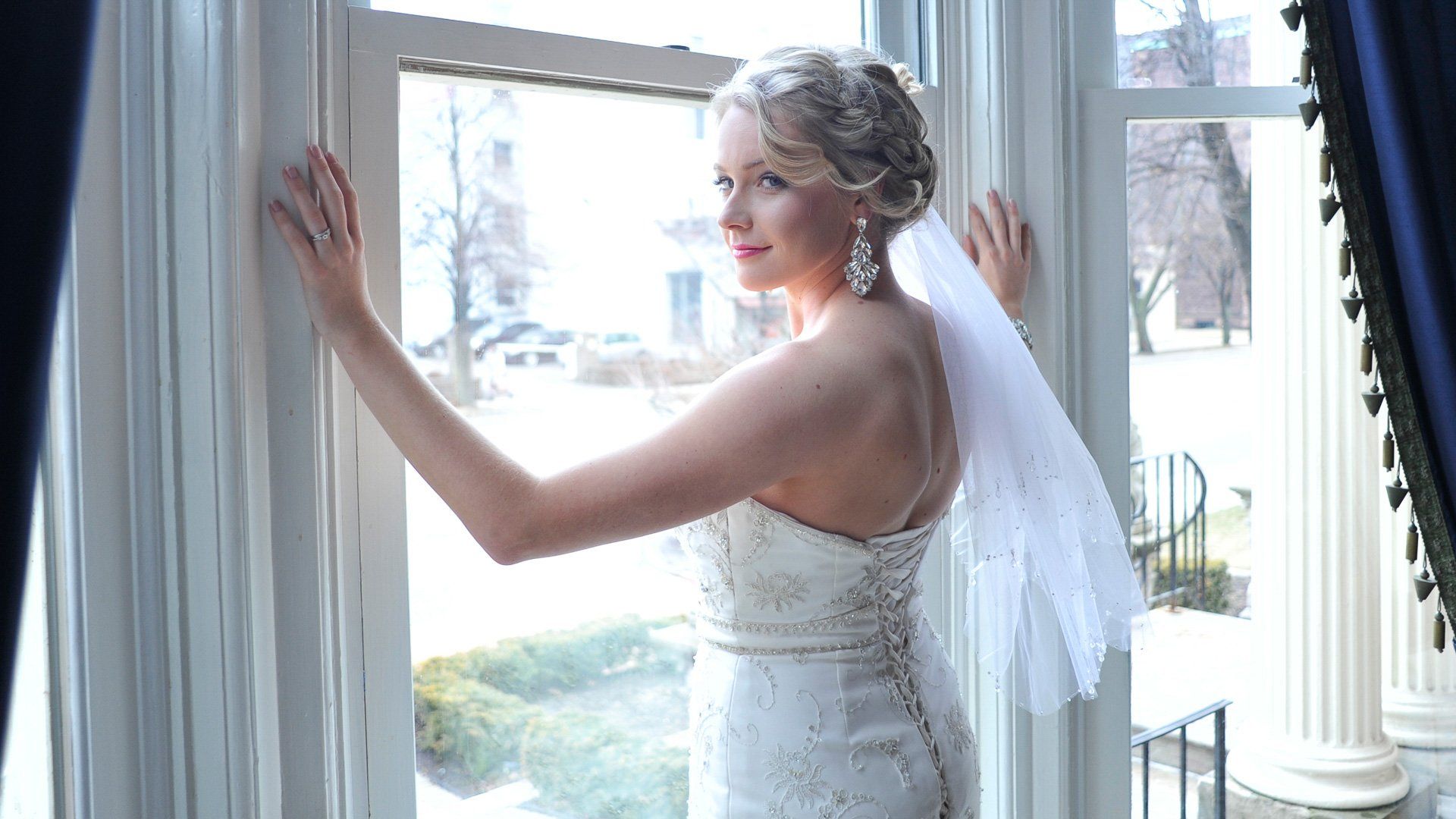 A woman in a wedding dress and veil is standing in front of a window.