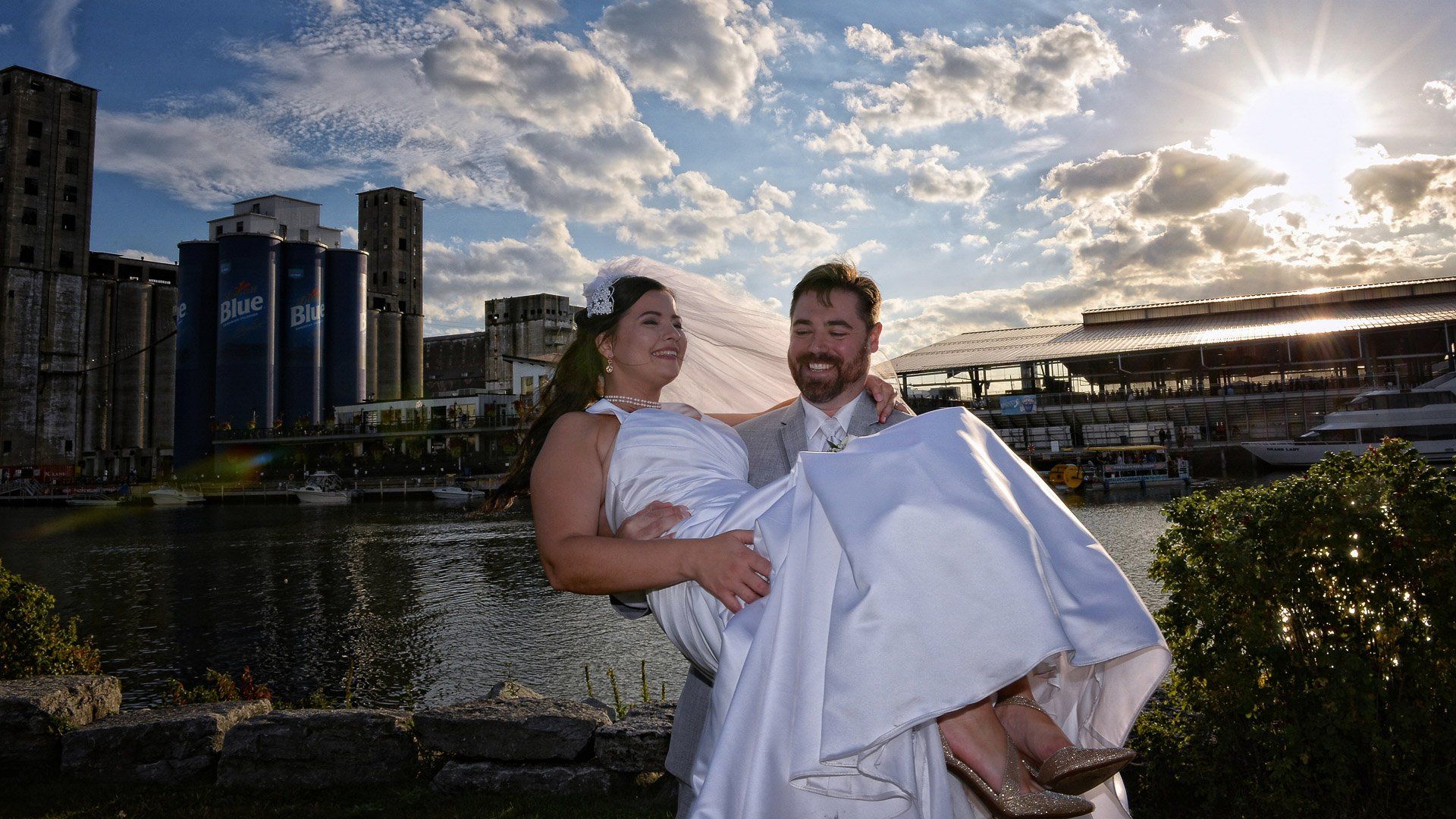 A man is carrying a bride in his arms in front of a body of water.