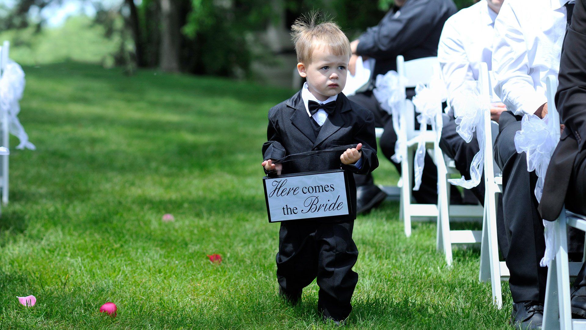 A ring bearer is walking down the aisle at a wedding holding a sign that says welcome to the bride.