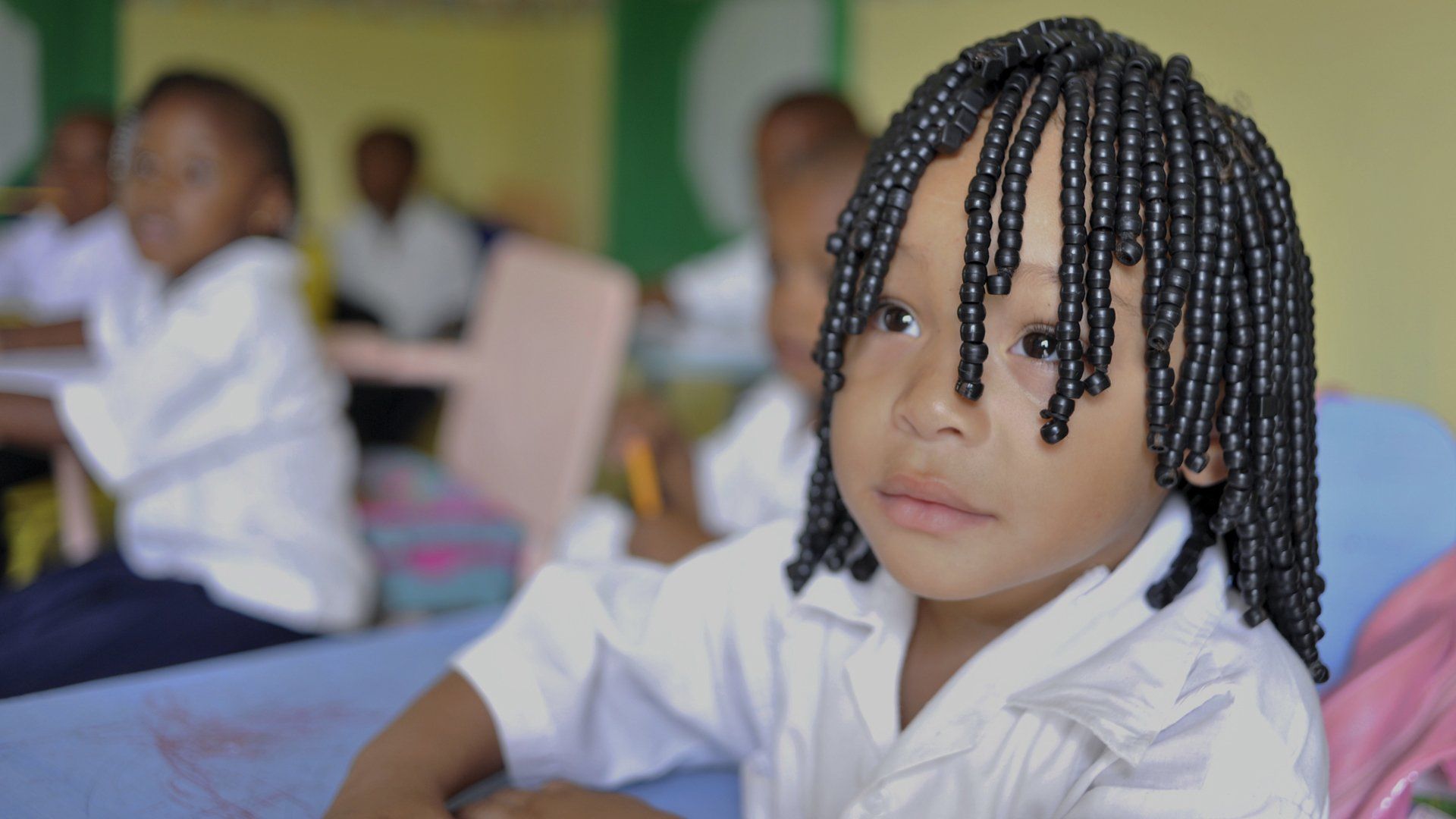 A young girl with dreadlocks is sitting at a desk in a classroom