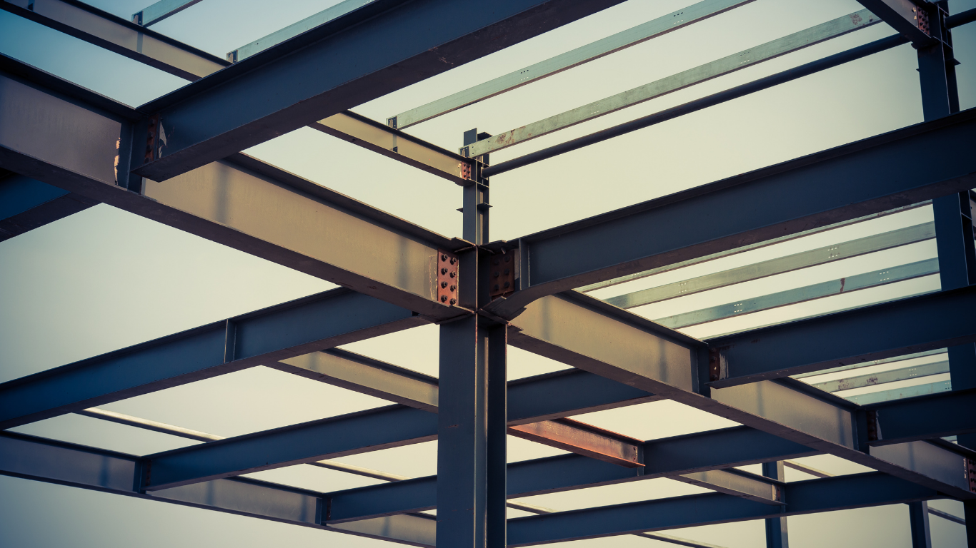 A person in work gloves using a drill press to create a hole in a metal beam, metal shavings visible.