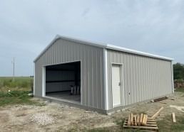 Gray metal building with a large open garage door and a side door in a rural setting.