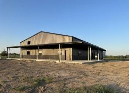 Metal building with a covered porch on a plot of land under a blue sky.