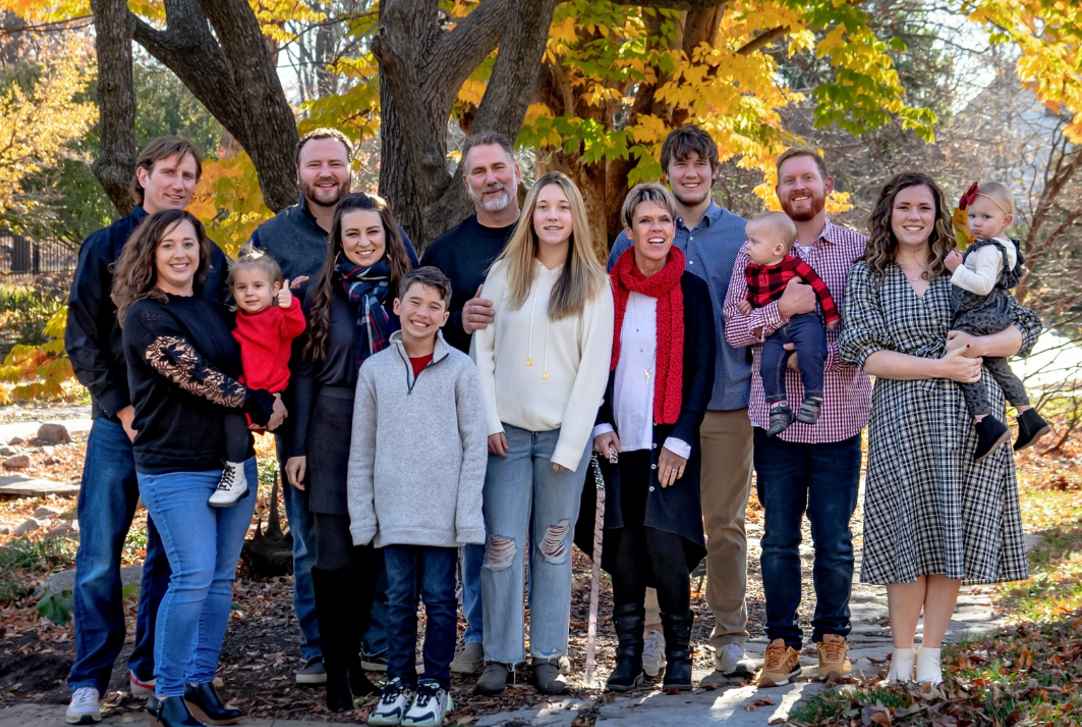 Large family posing outdoors in fall, smiling, with a mix of adults, children, and babies.
