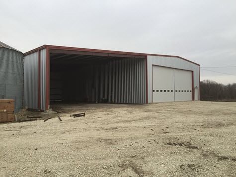 Metal shed with a large opening and a closed garage door, on a gravel lot, overcast sky.