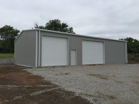 Gray metal building with two garage doors and gravel driveway under cloudy sky.