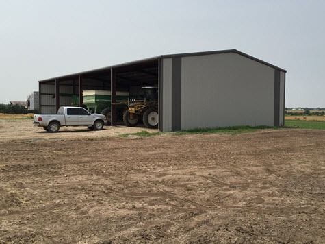 A gray farm shed with a tractor, a grain cart, and a white pickup truck on a dirt lot under a blue sky.