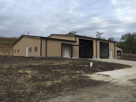 Tan metal building with two large garage doors, white doors, and a paved driveway, set on a dry, brown landscape.