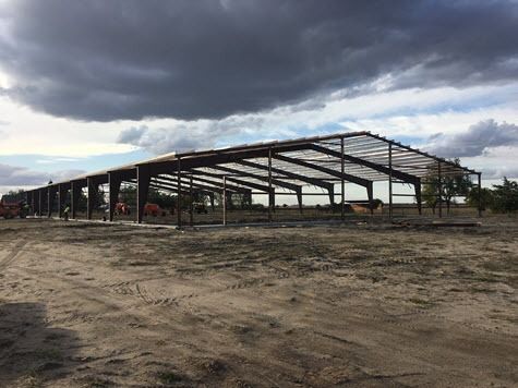 Steel frame of a large building under construction on a dirt lot, cloudy sky overhead.