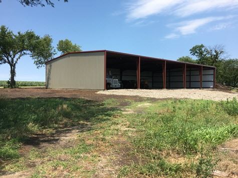 Metal agricultural building with open bays on a gravel area, tractor inside. Green field, trees, and blue sky.