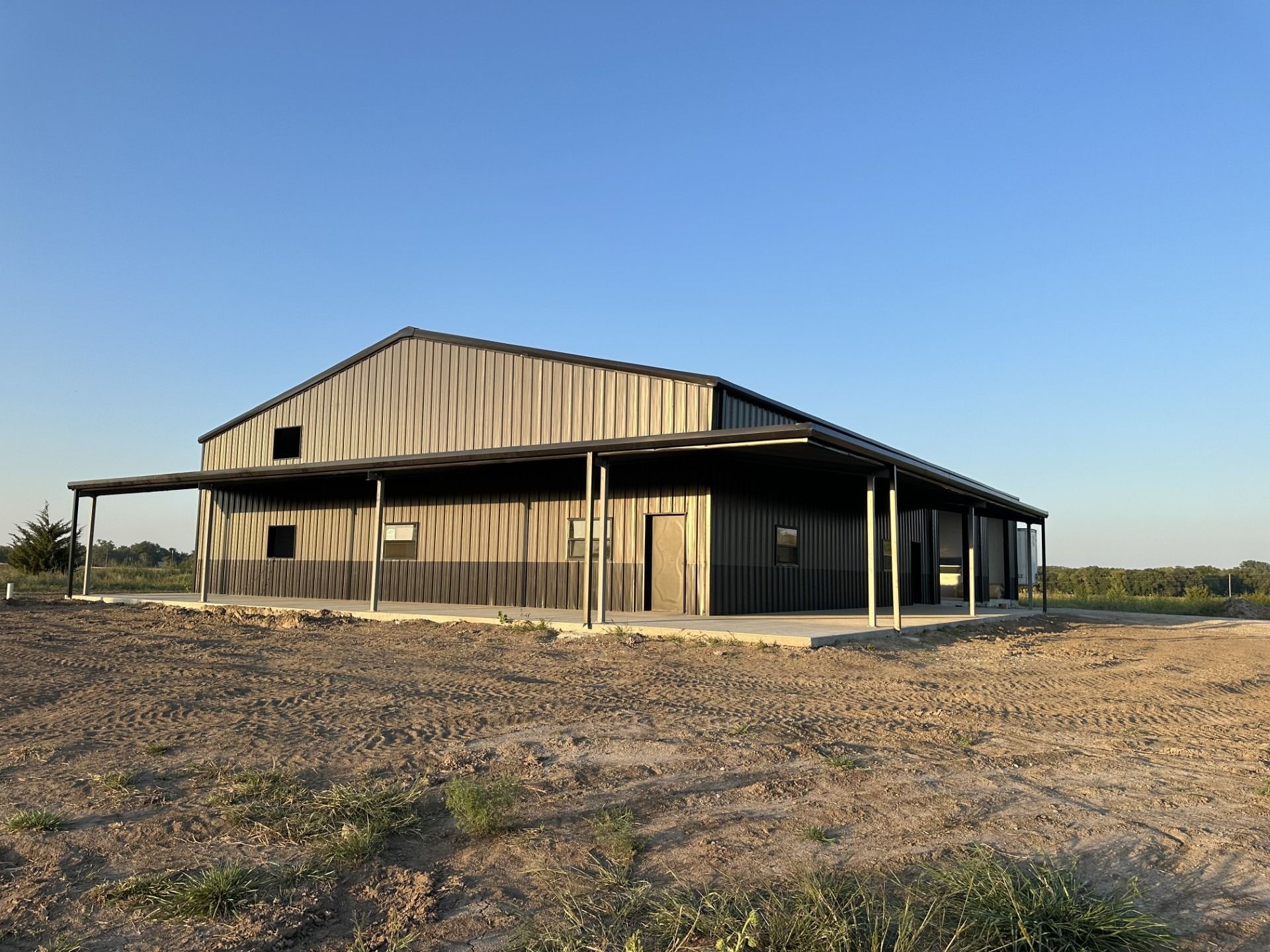 A large, gray metal barn with a wrap-around covered porch sits on a barren, brown field under a blue sky.