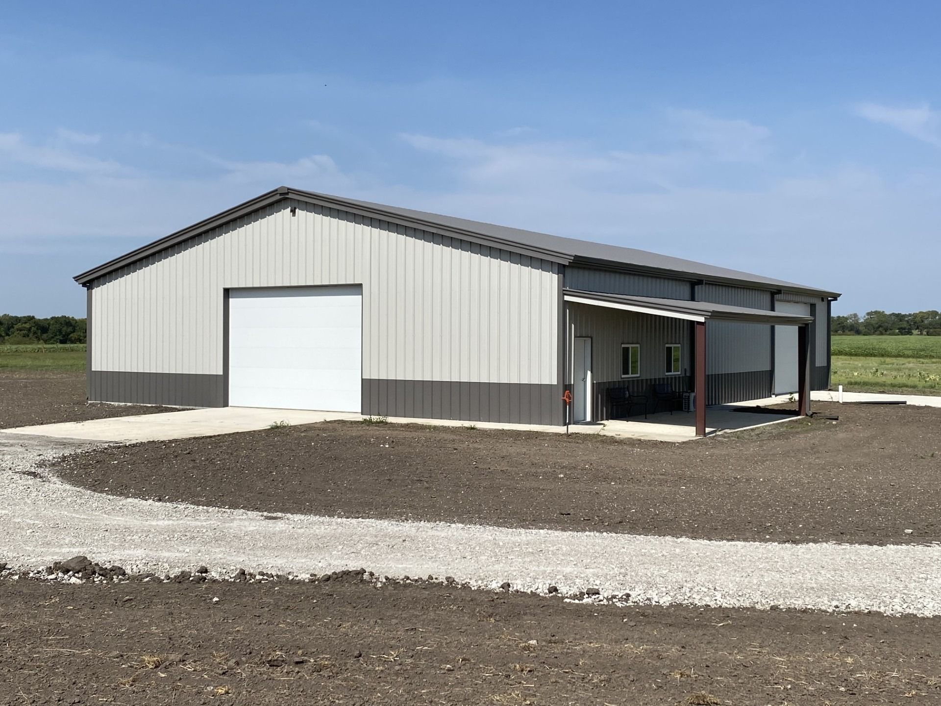 Steel building with white and dark gray siding, a white garage door, and a gravel driveway under a blue sky.