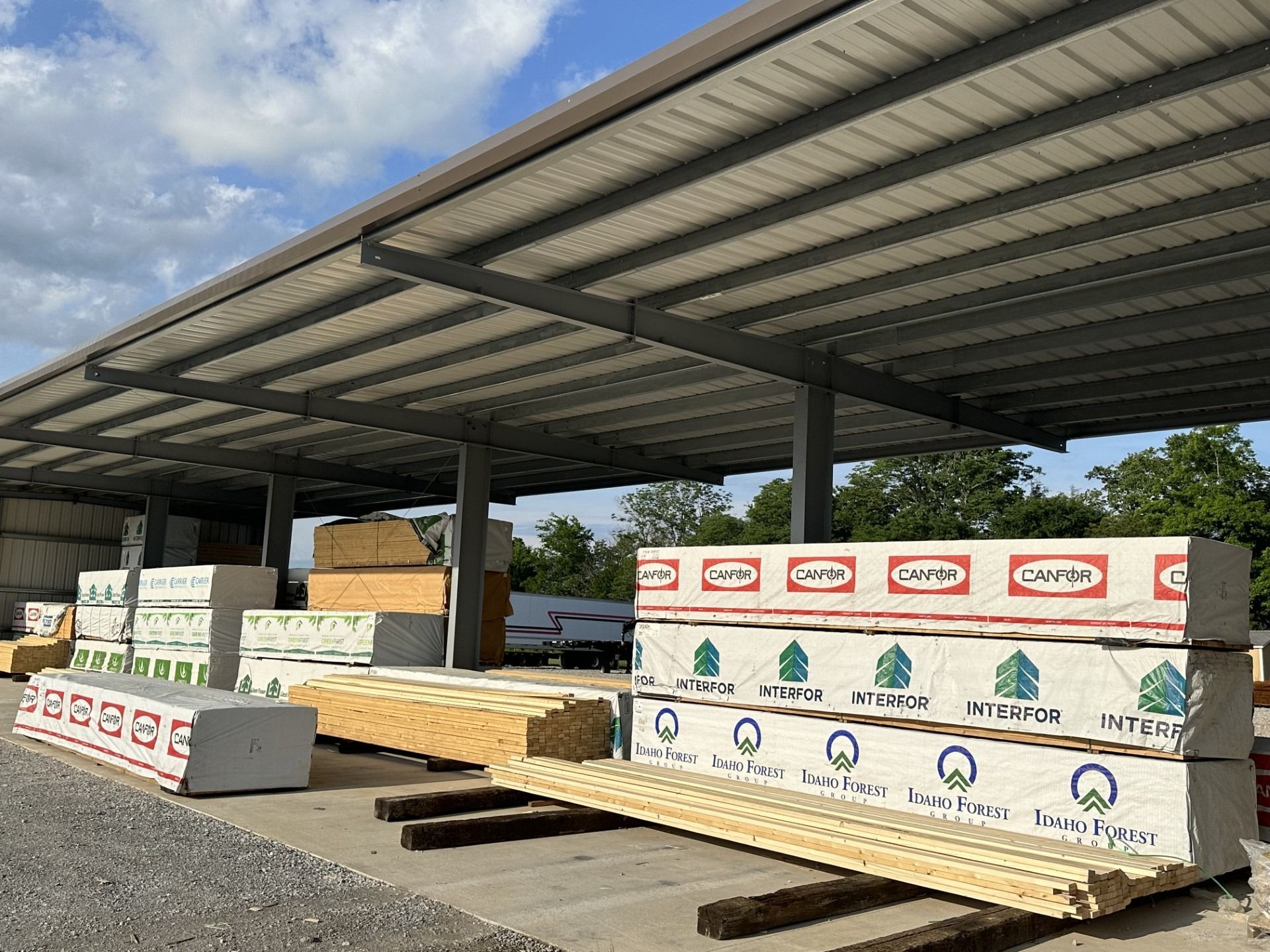 Lumber stacked under a metal awning in a lumberyard on a partly cloudy day.