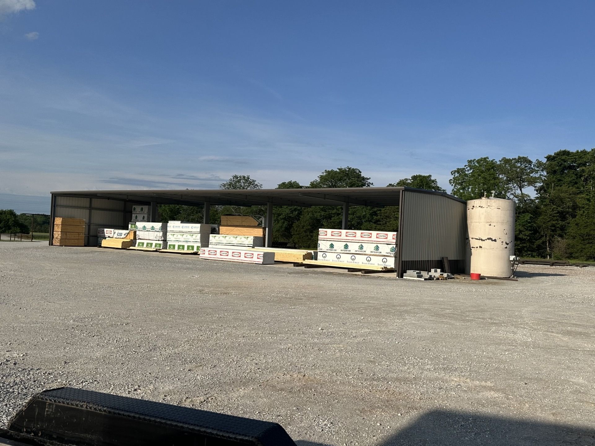 Piles of construction materials under a metal shelter on a gravel lot; a tall cylindrical tank is on the right.