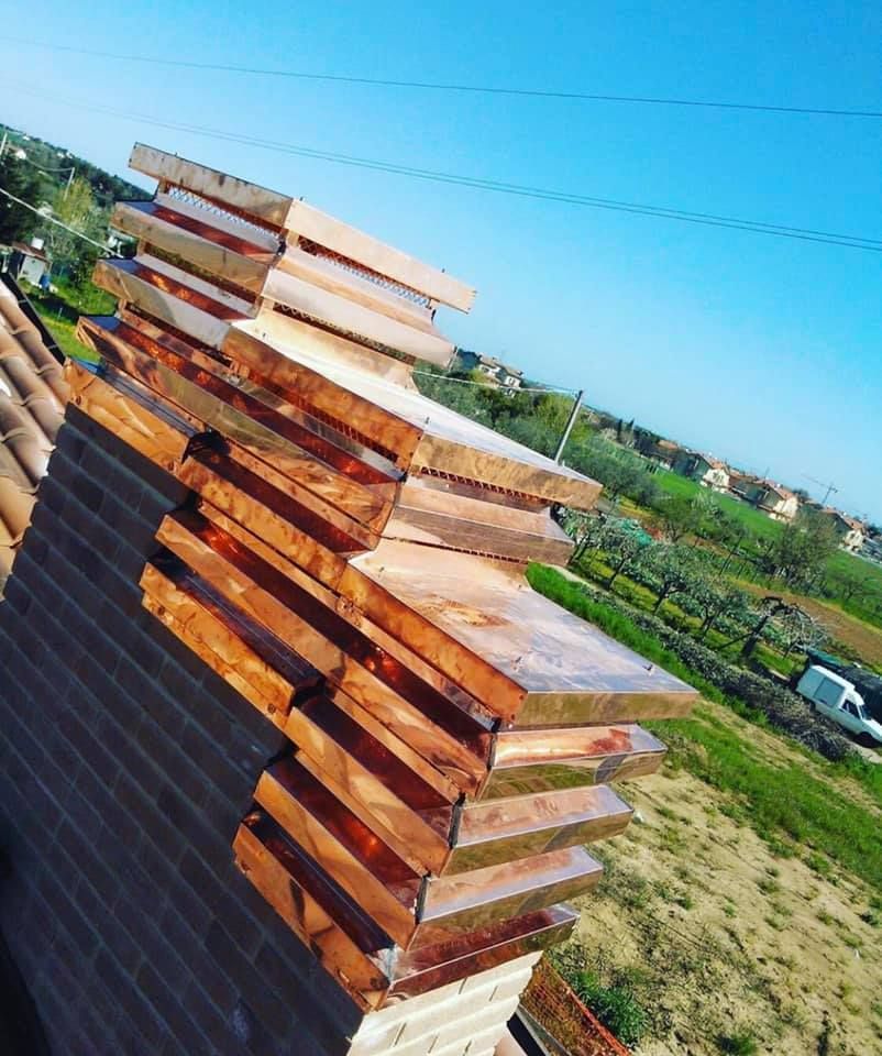 Cappello del comignolo in rame su un camino in mattoni contro un cielo azzurro, vicino a un campo.