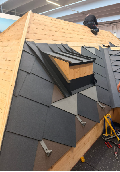 Operaio edile che installa tegole grigio scuro su un tetto con struttura in legno e lucernario.