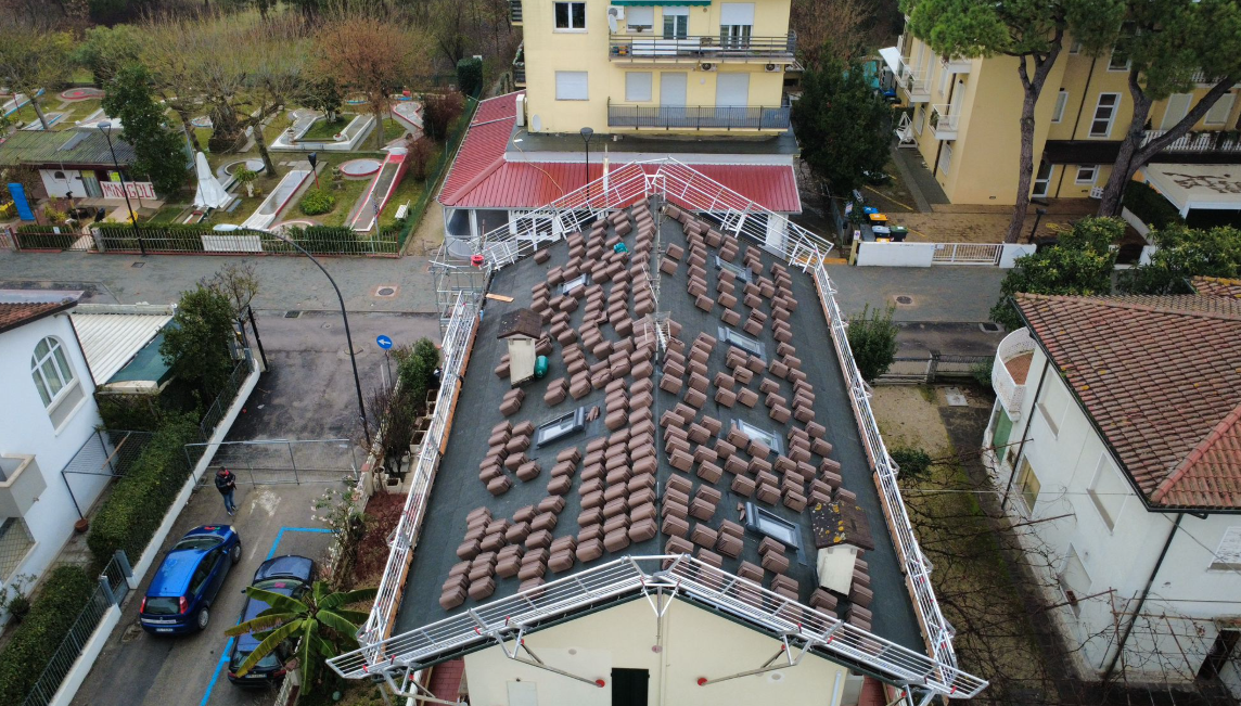 Tetto dell'edificio parzialmente ricoperto di tegole di terracotta; altre tegole sono sparse. Sono visibili la strada e gli edifici vicini.