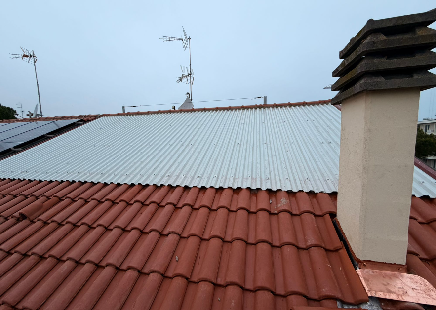 Vista di un tetto con tegole rosse e lamiera ondulata, camino e antenne in una giornata nuvolosa.