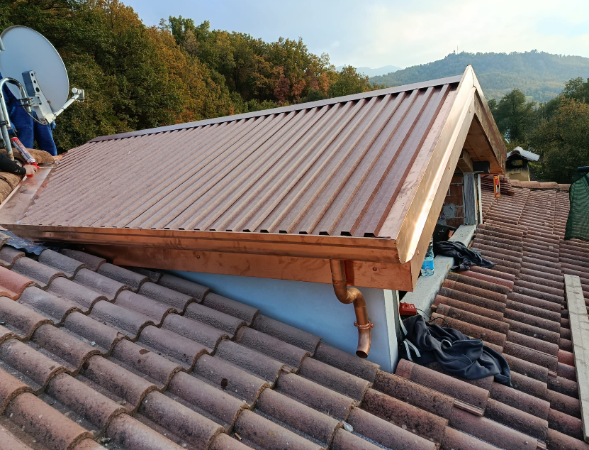 Tetto in metallo color rame di un edificio, con tetto in tegole di terracotta in primo piano; all'esterno, alberi sullo sfondo.