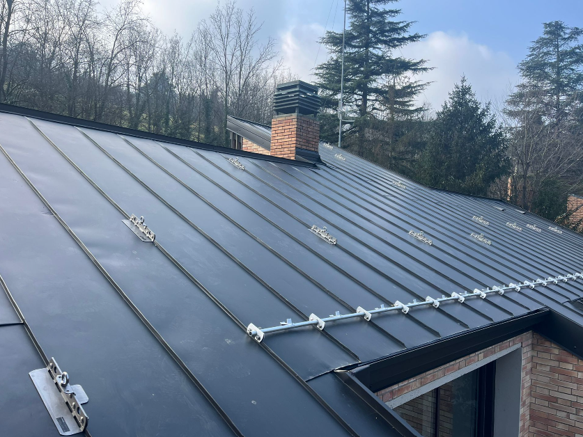 Tetto in metallo nero con imbracatura di sicurezza, camino e alberi sullo sfondo.