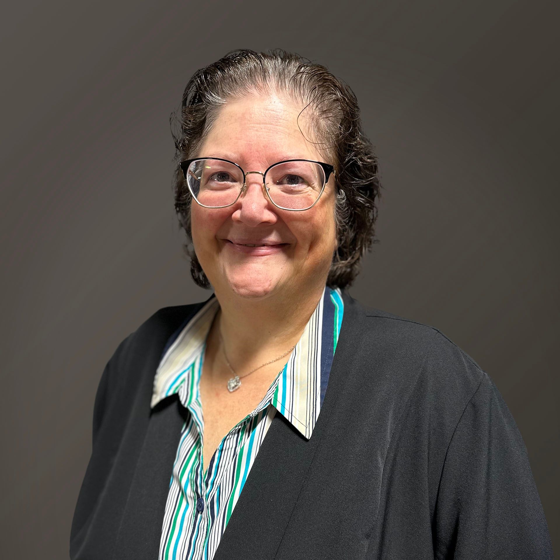 Woman wearing glasses, smiling, with dark hair, a patterned shirt and a black jacket.