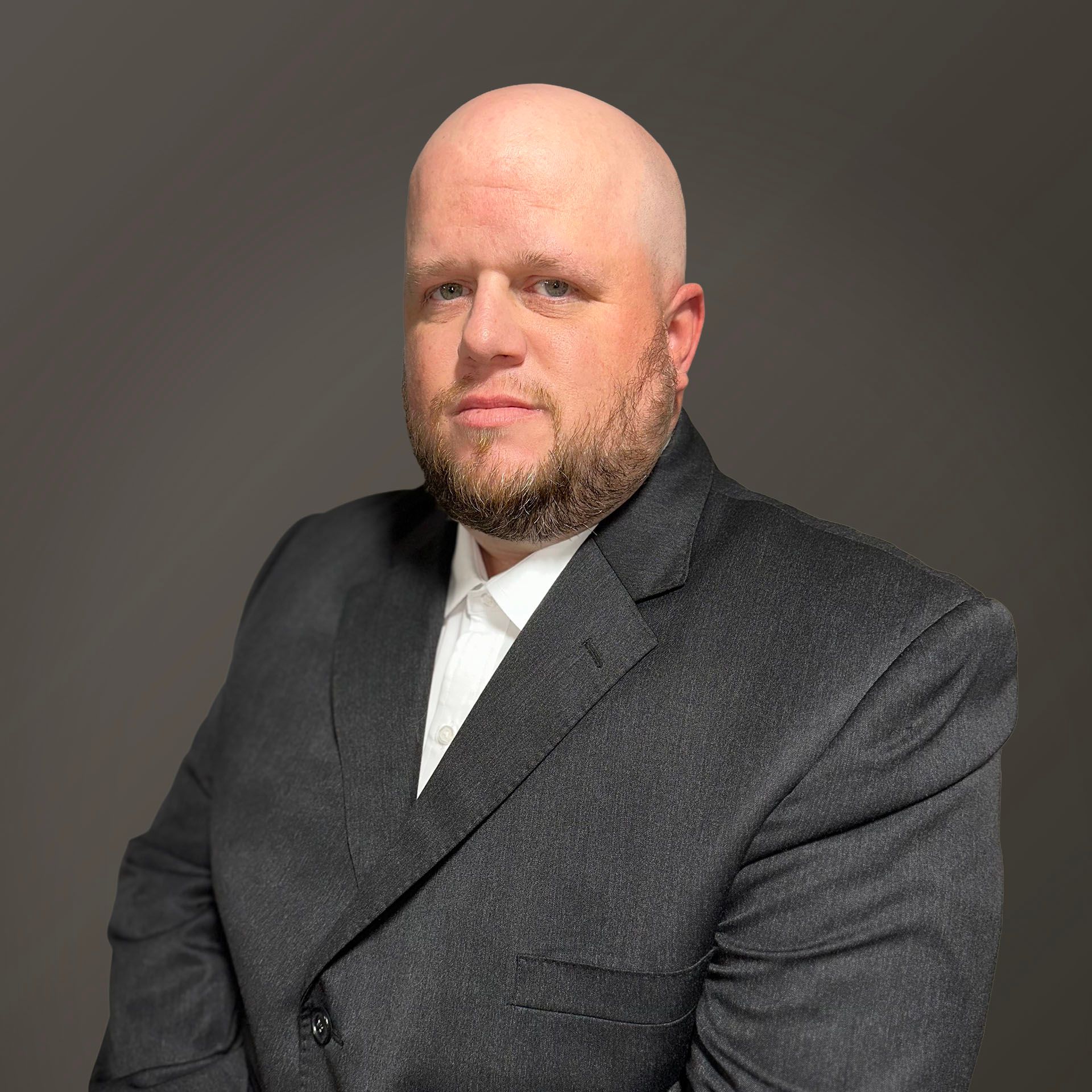 Bald man with a beard, wearing a black suit and white shirt, posing against a gray background.