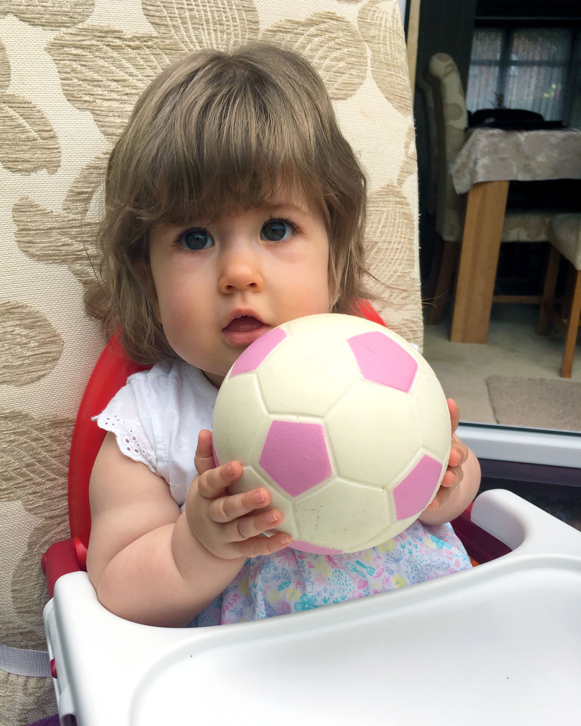 A baby girl is sitting in a high chair holding a pink and white soccer ball