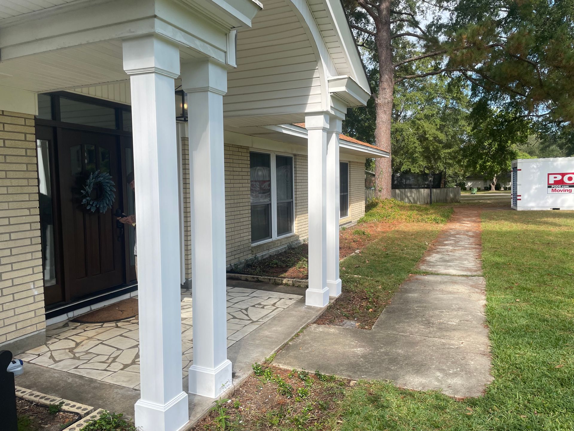 White-columned porch of a brick house with a walkway; a For Sale moving container is in the background.