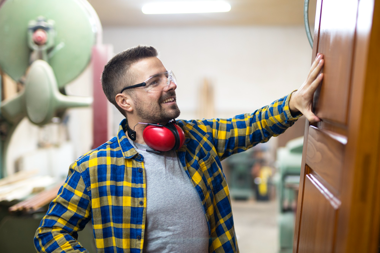 “Carpenter inspecting a finished wooden door in a workshop while wearing safety glasses and ear prot