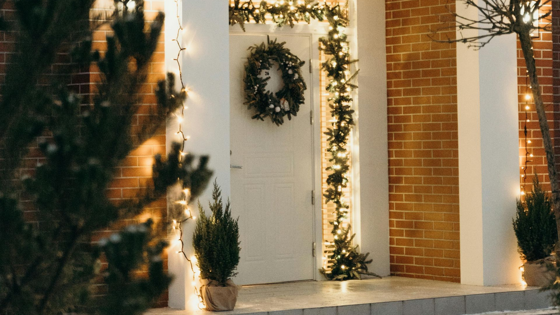 A white front door decorated with a wreath and illuminated evergreen garlands against a brick house exterior.