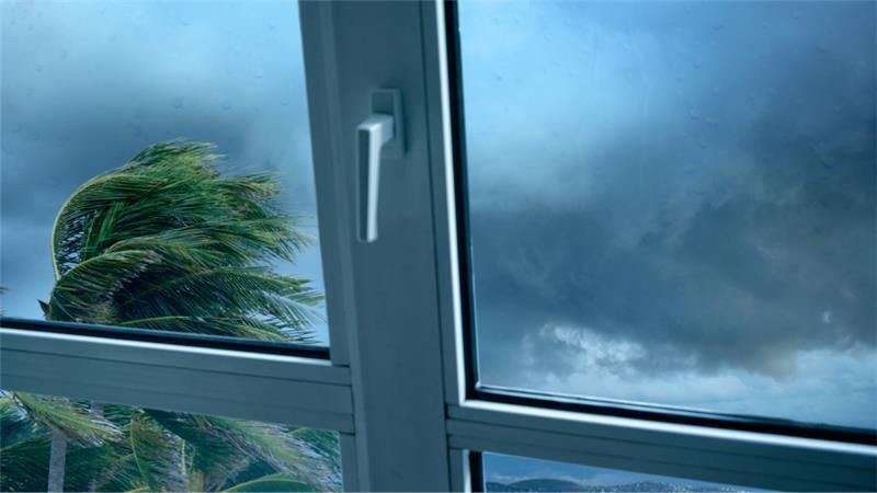 “View through a closed window during a storm with strong winds bending palm trees and dark clouds ou