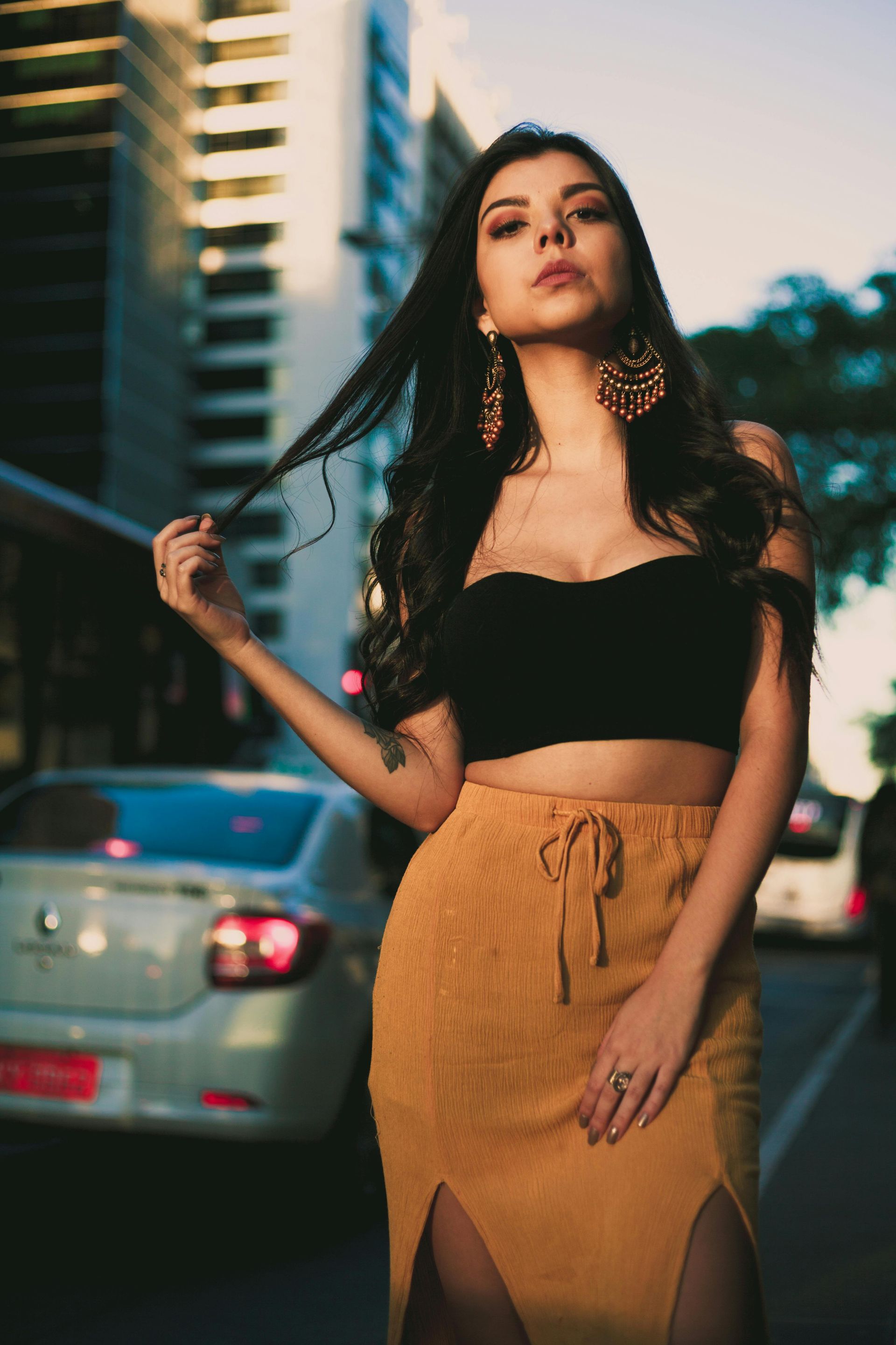 Woman in black crop top and yellow skirt, touching hair, standing on a city street.