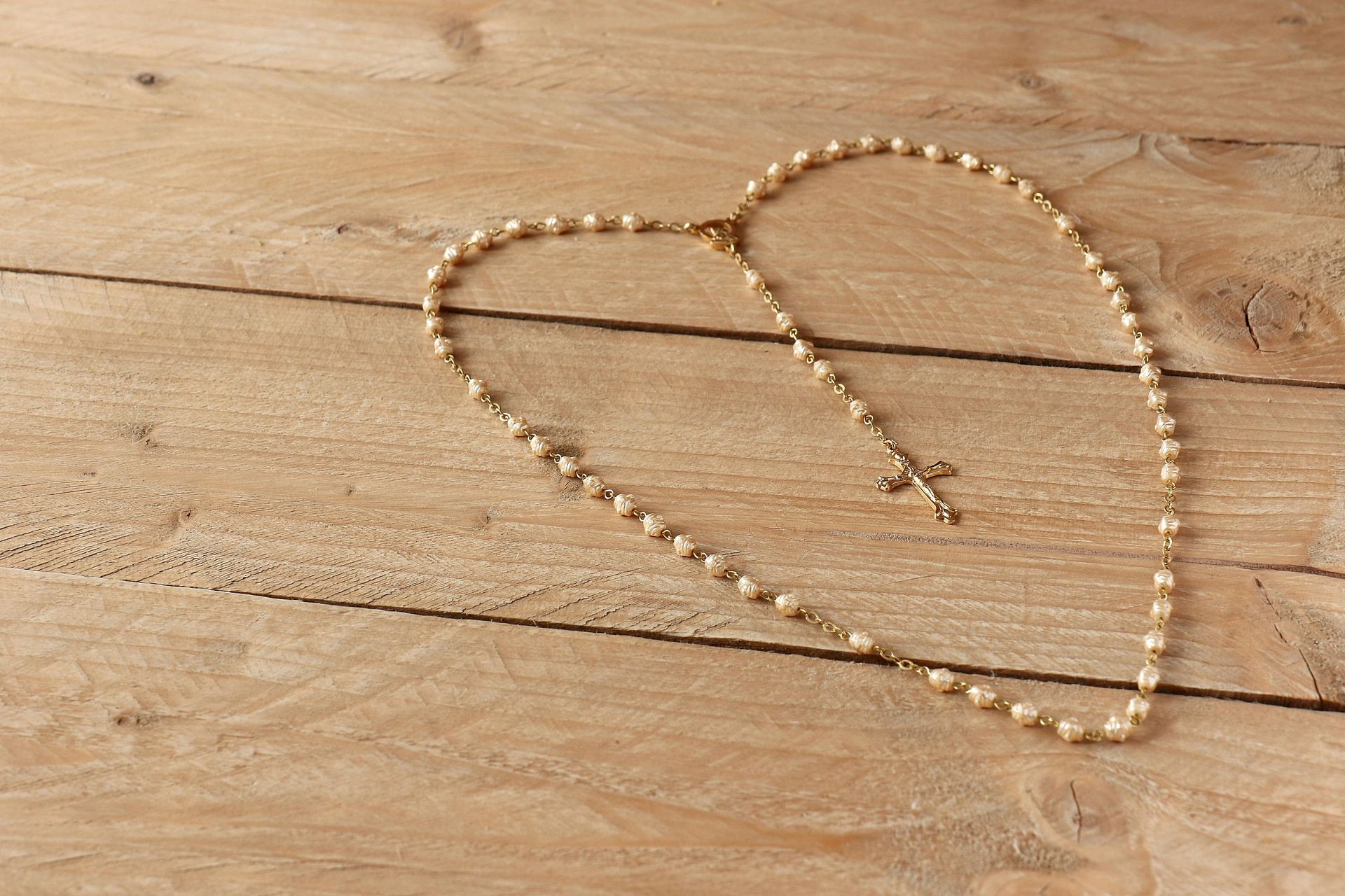 Rosary necklace, gold beads, and cross arranged in a heart shape on a light wood surface.