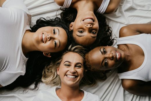 Four diverse women smiling, heads together, on white bedding.