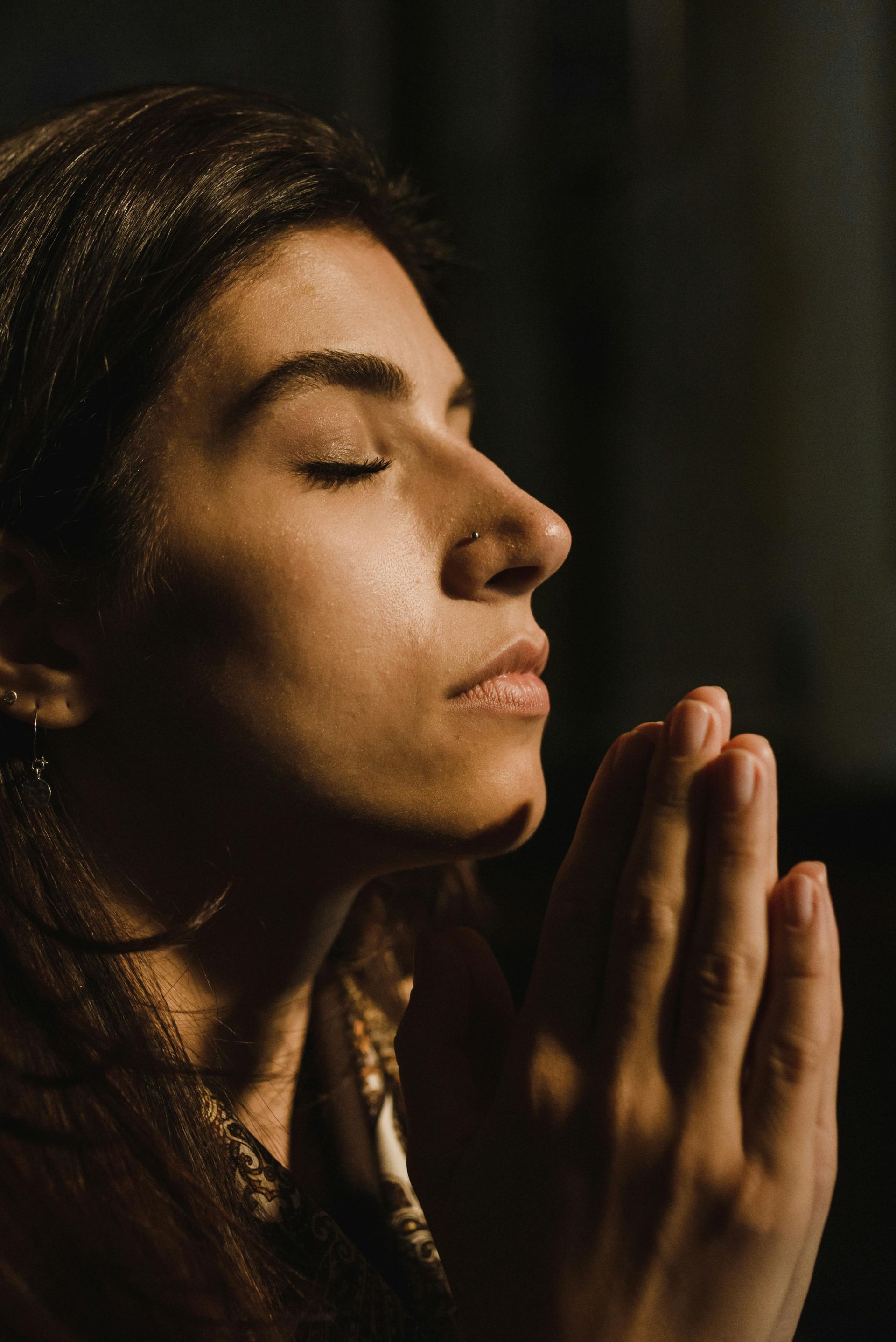 Woman with eyes closed, hands clasped in prayer, illuminated by warm light, dark background.