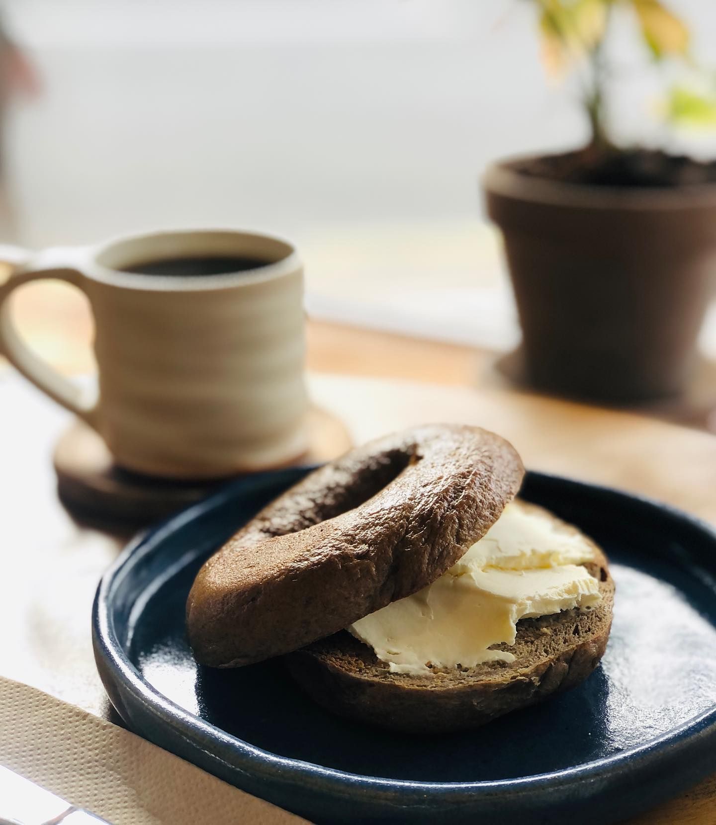 A bagel with butter on it is on a plate next to a cup of coffee