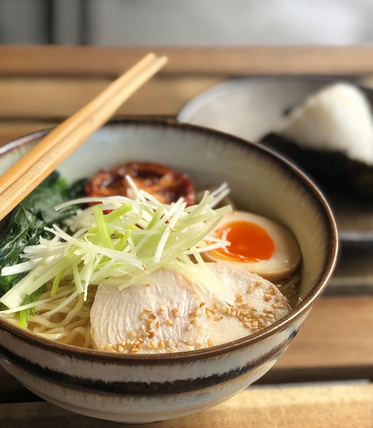 A bowl of ramen with noodles , chicken , egg and green onions on a table with chopsticks.