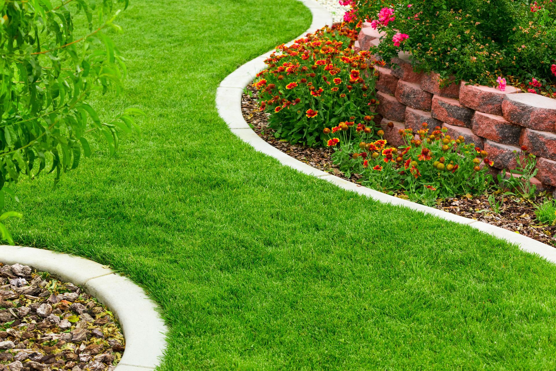 Curved garden path bordered by bright green grass, flowers, and a low brick retaining wall
