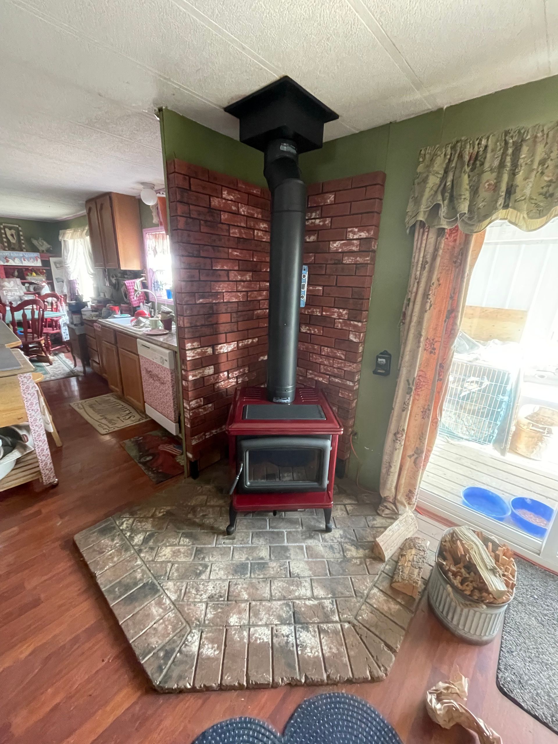 Red wood-burning stove with black chimney pipe, set on a brick hearth and backed by a brick wall.