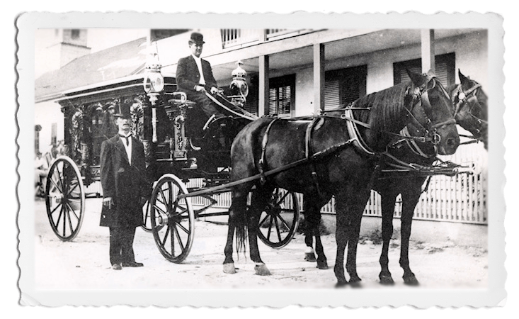 A black-and-white photo shows a horse-drawn funeral hearse with a driver, standing beside a man in a formal suit.