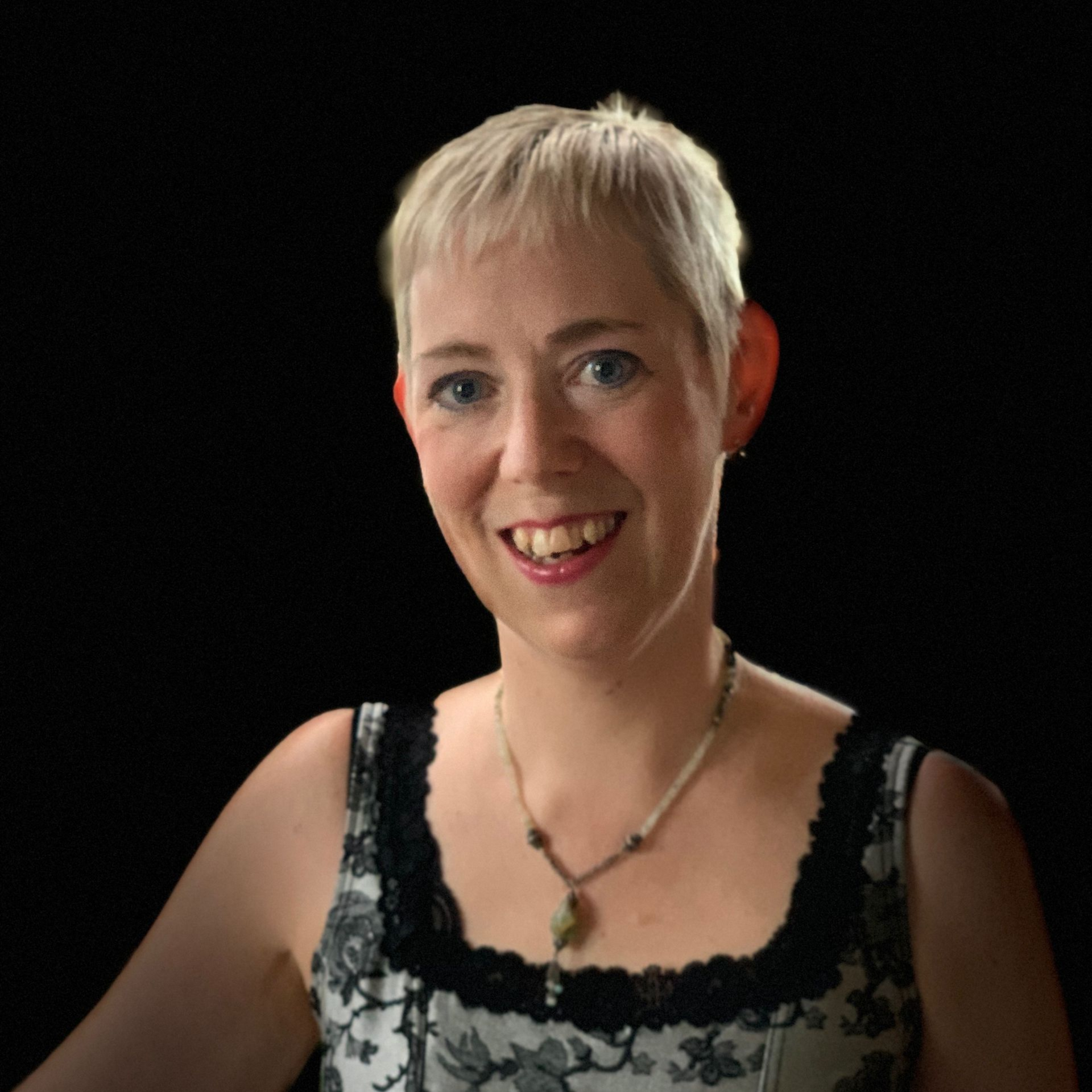 Woman (Christina Laberge) with short blonde hair smiles, wearing a patterned top and necklace against a black background.